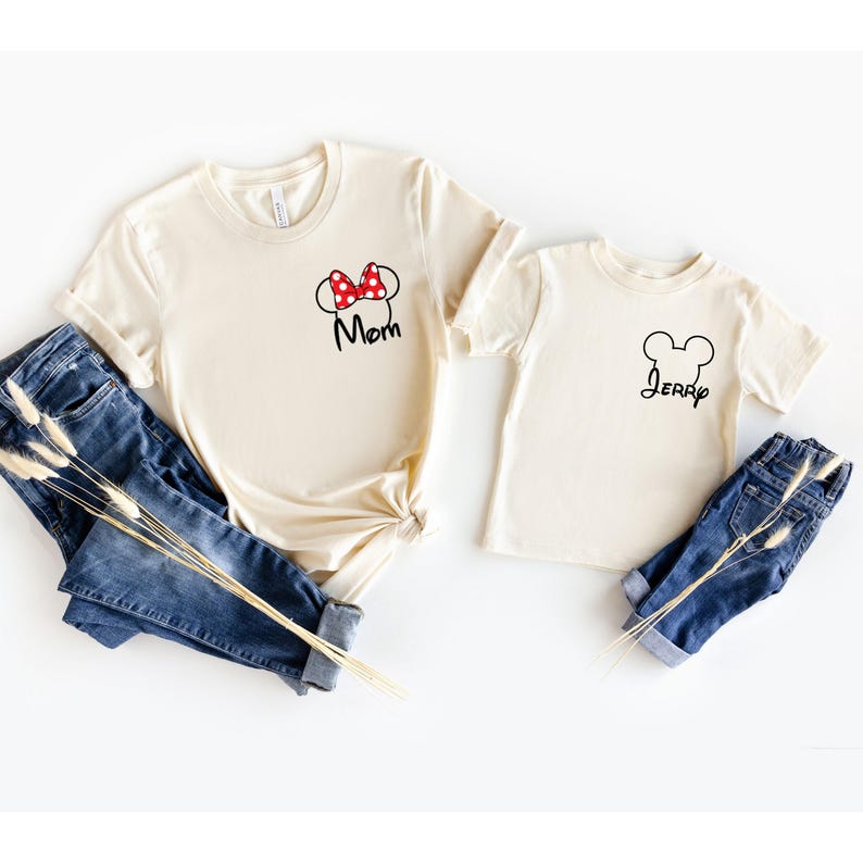 May include: Two cream-colored t-shirts with black text and graphics. One shirt has a Minnie Mouse bow and the word "Mom". The other shirt has a Mickey Mouse head outline and the name "Jerry". Blue jeans are next to the shirts.