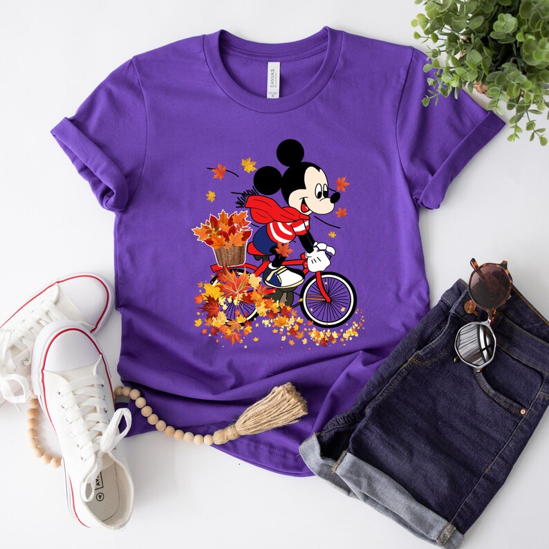 May include: A purple t-shirt featuring a graphic of Mickey Mouse riding a red bicycle through autumn leaves. Mickey is wearing a red scarf and carrying a basket of leaves. White sneakers and denim shorts are also in the image.
