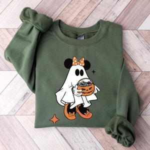 May include: An olive green sweatshirt featuring a Halloween-themed graphic. The graphic depicts a cartoon ghost with mouse ears, an orange bow, and holding a jack-o'-lantern filled with candy. The design includes orange and black accents, and a star and diamond shape.