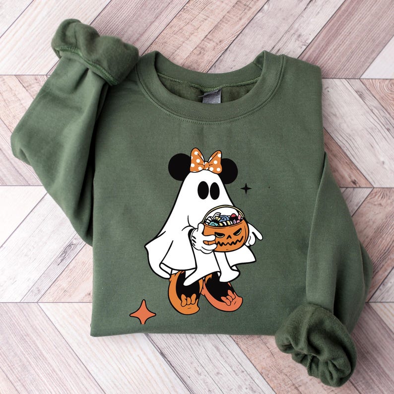 May include: An olive green sweatshirt featuring a Halloween-themed graphic. The graphic depicts a cartoon ghost with mouse ears, an orange bow, and holding a jack-o'-lantern filled with candy. The design includes orange and black accents, and a star and diamond shape.