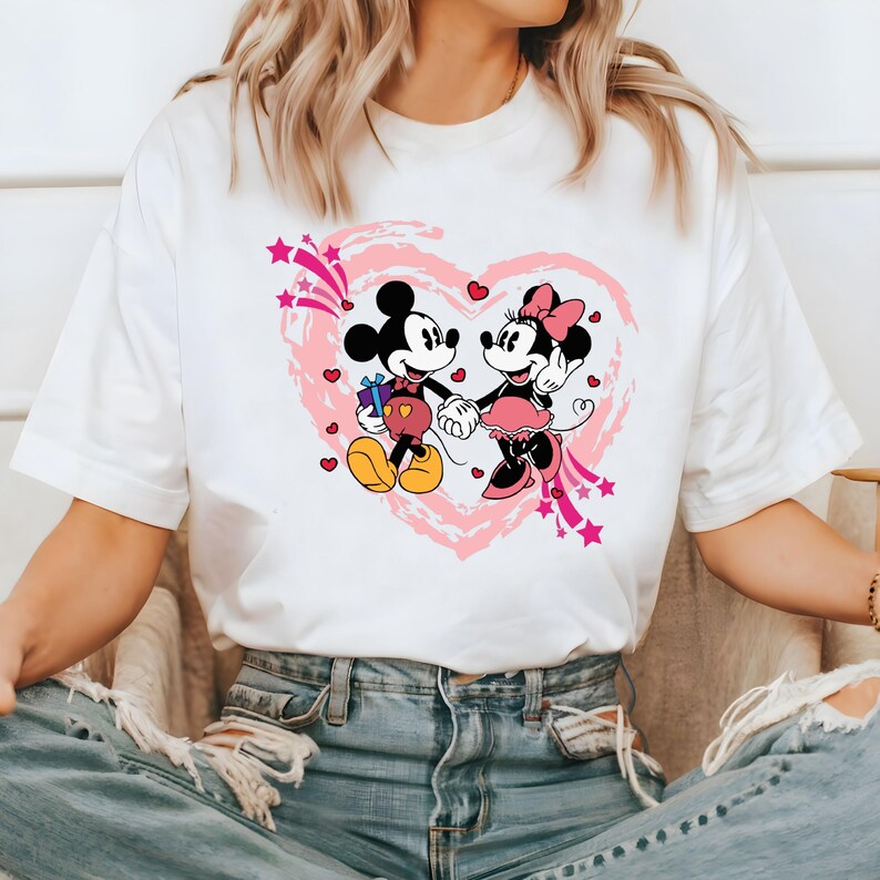 May include: White t-shirt featuring a graphic of Mickey and Minnie Mouse holding hands inside a pink heart. Mickey Mouse is holding a gift. The design includes pink stars and hearts, creating a romantic theme.