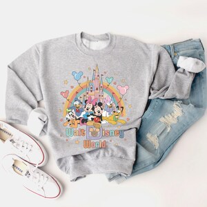 Puede incluir: Una sudadera gris con un gr&aacute;fico colorido de Mickey Mouse y sus amigos frente al Castillo de Cenicienta. El gr&aacute;fico tambi&eacute;n incluye un arco&iacute;ris y el texto "Walt Disney World".