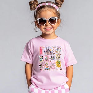 May include: A light pink t-shirt featuring a colorful Easter-themed graphic with Disney characters and the word "Happy". The shirt is paired with a pink and white checkered headband and shorts. The child is wearing white sunglasses.