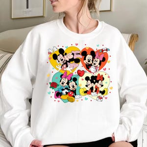 May include: White sweatshirt with a colorful graphic print of Mickey Mouse and Minnie Mouse in various poses. The graphic features hearts, stars, and other decorative elements.