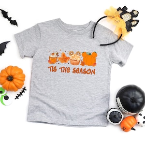 Pu&ograve; includere: T-shirt grigio a maniche corte con la scritta arancione "Tis the Season". La grafica include un pumpkin spice latte, foglie autunnali, un cupcake e zucche a forma di Mickey Mouse. Decorazioni di Halloween intorno.
