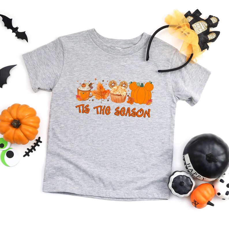 Pu&ograve; includere: T-shirt grigio a maniche corte con la scritta arancione "Tis the Season". La grafica include un pumpkin spice latte, foglie autunnali, un cupcake e zucche a forma di Mickey Mouse. Decorazioni di Halloween intorno.