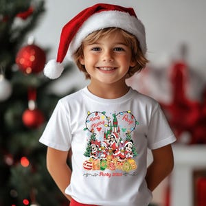 May include: A child wearing a white t-shirt with a colorful Christmas graphic featuring Disney characters and the text "Very Merry Christmas Party 2025." The child is also wearing a red and white Santa hat.