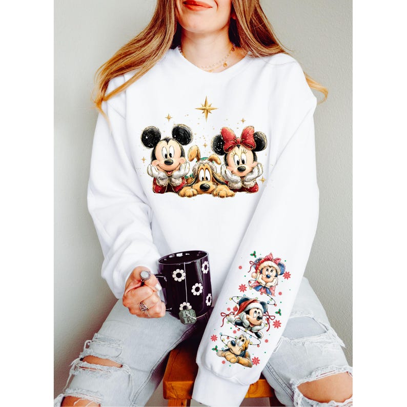 May include: White sweatshirt featuring Mickey Mouse, Minnie Mouse, and Pluto in festive attire. The sleeve displays additional Disney characters in Christmas decorations. A black mug with white floral designs is held in the hand.