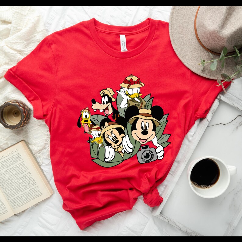 May include: Red t-shirt featuring a graphic print of Mickey Mouse, Minnie Mouse, Goofy, Donald Duck, and Pluto dressed in safari gear. The characters are surrounded by green leaves and Mickey Mouse is holding a camera.