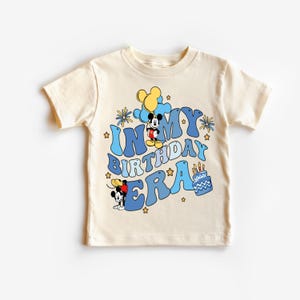 May include: A light beige t-shirt with a blue and yellow graphic design featuring Mickey Mouse and the text "In My Birthday Era".