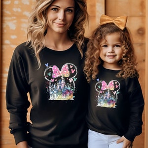 Op de afbeelding: Twee mensen dragen zwarte sweatshirts met een kleurrijke afbeelding van Assepoester's kasteel in een Mickey Mouse-hoofd met een roze strik. De afbeelding bevat ook vuurwerk en Mickey Mouse-oren.