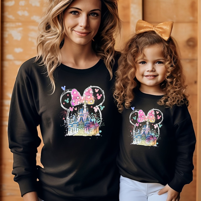 Op de afbeelding: Twee mensen dragen zwarte sweatshirts met een kleurrijke afbeelding van Assepoester's kasteel in een Mickey Mouse-hoofd met een roze strik. De afbeelding bevat ook vuurwerk en Mickey Mouse-oren.