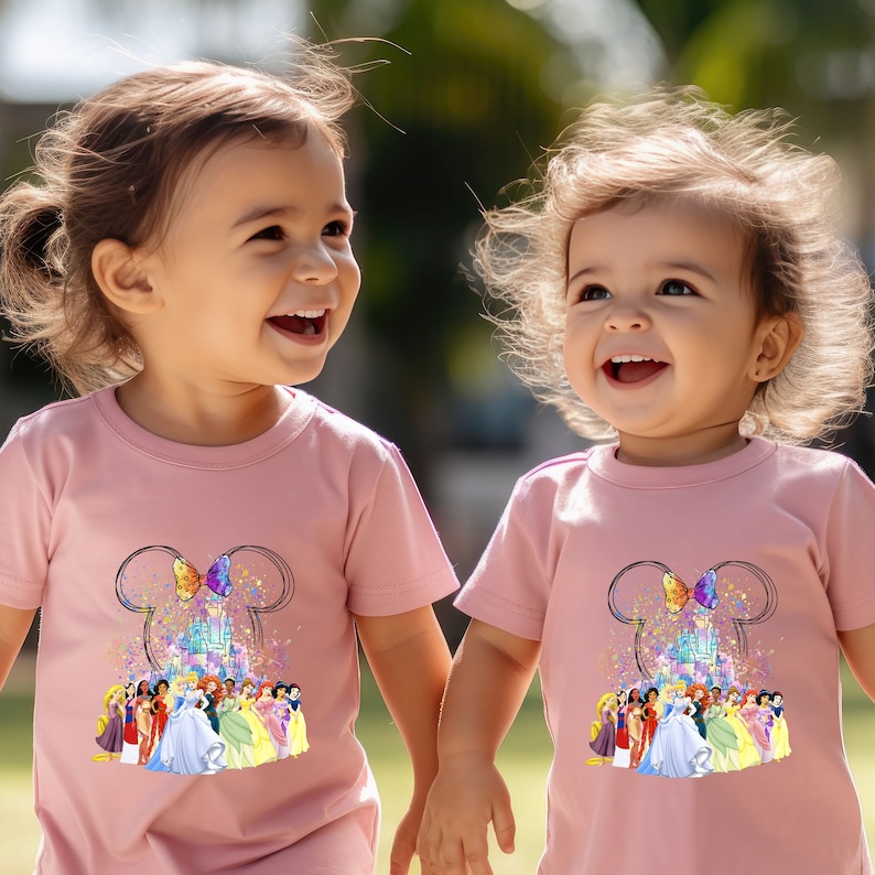 Puede incluir: Dos ni&ntilde;as peque&ntilde;as con camisetas rosas con un gr&aacute;fico de princesas Disney y el castillo de Cenicienta. Las camisetas tienen un dise&ntilde;o de orejas de Mickey con un lazo.