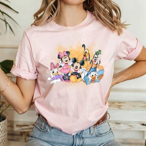 Puede incluir: Una camiseta rosa con un estampado gr&aacute;fico de Mickey Mouse, Minnie Mouse, Donald Duck, Goofy y Pluto. Los personajes est&aacute;n todos sonriendo y parecen estar divirti&eacute;ndose juntos.