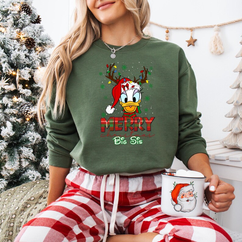 May include: Green Christmas sweatshirt featuring Donald Duck wearing a Santa hat and reindeer antlers with string lights. The words "MERRY" and "Big Sis" are printed below the graphic. The person is wearing red and white plaid pajama pants and holding a mug with a Santa Claus design.