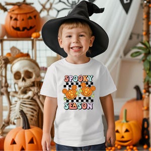 May include: A child wearing a black witch hat and a white t-shirt with the words "Spooky Season" and two orange pumpkin designs. The background includes Halloween decorations such as pumpkins and a skeleton.