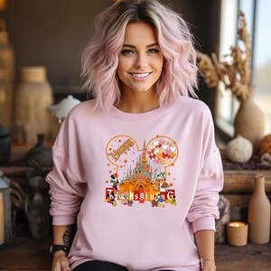 May include: A light pink sweatshirt featuring a graphic of a castle, Mickey Mouse, Minnie Mouse, and other Disney characters. The word "Happy" is above the castle, and "Thanksgiving" is below. The design includes balloons and fall leaves.