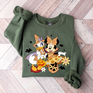 May include: Green sweatshirt with a graphic print of Minnie Mouse and Daisy Duck dressed in Halloween costumes. Minnie Mouse is wearing a brown witch costume with a pumpkin skirt and a witch hat. Daisy Duck is wearing a purple witch costume with a star on her hat. They are both holding a pumpkin bucket.