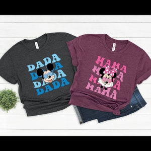 May include: Two t-shirts with a retro design featuring Mickey Mouse and Minnie Mouse. The dark grey t-shirt has the word "DADA" repeated in blue and pink with Mickey Mouse wearing sunglasses. The maroon t-shirt has the word "MAMA" repeated in pink with Minnie Mouse wearing a pink bow and sunglasses.