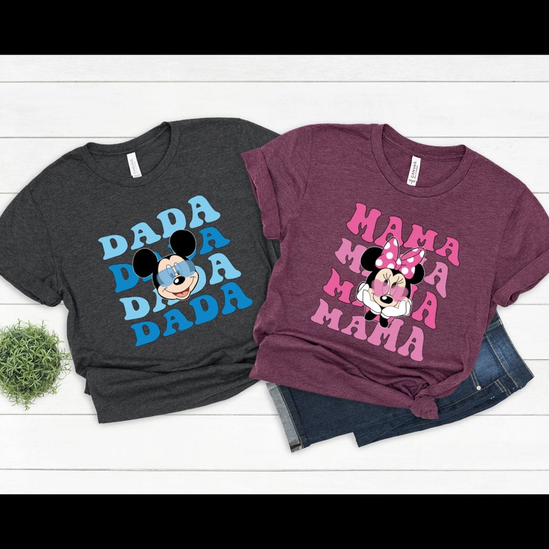 May include: Two t-shirts with a retro design featuring Mickey Mouse and Minnie Mouse. The dark grey t-shirt has the word "DADA" repeated in blue and pink with Mickey Mouse wearing sunglasses. The maroon t-shirt has the word "MAMA" repeated in pink with Minnie Mouse wearing a pink bow and sunglasses.