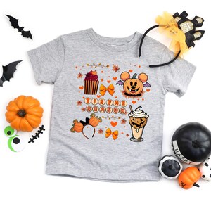 Puede incluir: Una camiseta gris de manga corta con un dise&ntilde;o tem&aacute;tico de Halloween. El dise&ntilde;o incluye una calabaza, una magdalena y una bebida de caf&eacute;, junto con el texto "Tis the Season". Tambi&eacute;n hay elementos decorativos como lazos y hojas.