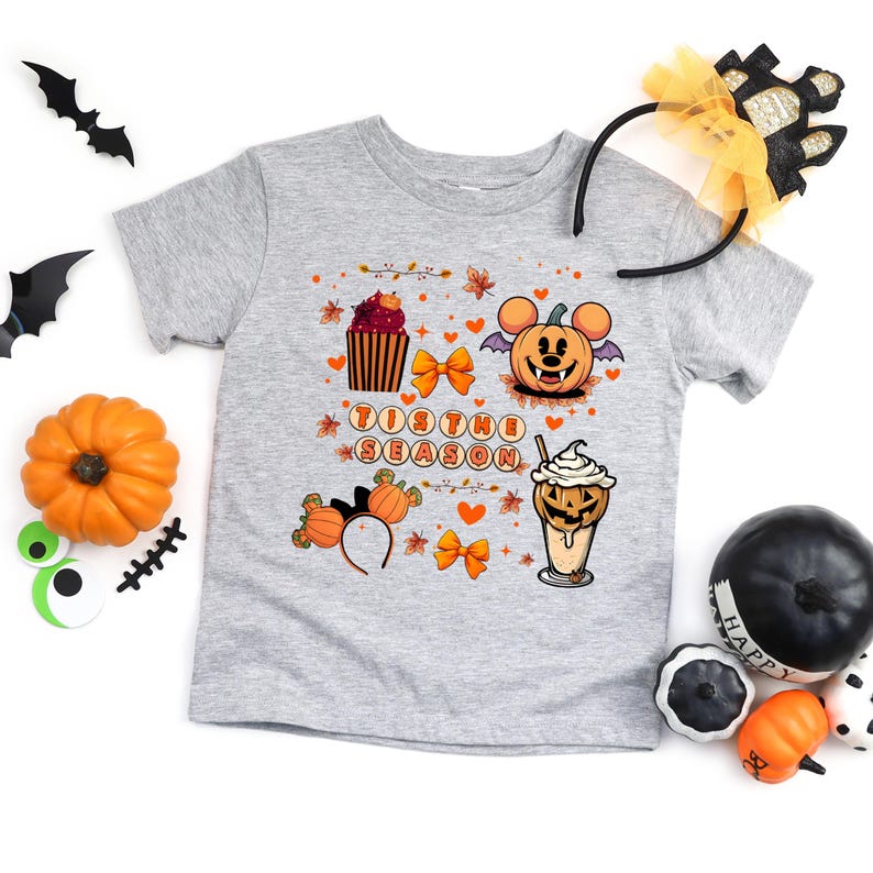 Puede incluir: Una camiseta gris de manga corta con un dise&ntilde;o tem&aacute;tico de Halloween. El dise&ntilde;o incluye una calabaza, una magdalena y una bebida de caf&eacute;, junto con el texto "Tis the Season". Tambi&eacute;n hay elementos decorativos como lazos y hojas.