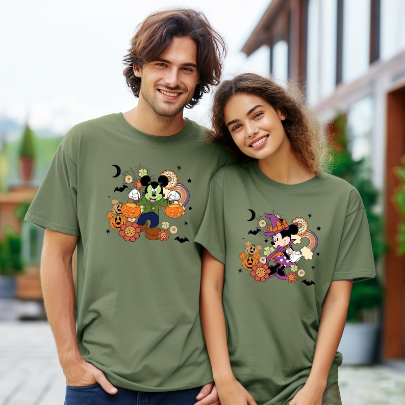 K&ouml;nnte beinhalten: Zwei Personen tragen olivgr&uuml;ne T-Shirts mit Halloween-Motiven von Mickey und Minnie Mouse. Mickey Mouse h&auml;lt K&uuml;rbisse, w&auml;hrend Minnie Mouse einen Hexenhut tr&auml;gt. Beide Designs enthalten Regenbogen, Blumen und Flederm&auml;use.