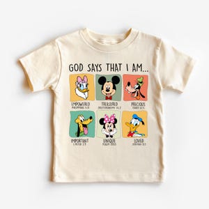 Puede incluir: Camiseta color crema con personajes de Disney y el texto "GOD SAYS THAT I AM...". La camiseta muestra a Daisy Duck, Mickey Mouse, Goofy, Pluto, Minnie Mouse y Donald Duck, cada uno con una afirmaci&oacute;n positiva.