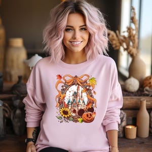 May include: A light pink sweatshirt featuring a colorful graphic of a castle, sunflowers, and a jack-o'-lantern. The graphic is framed by a decorative ribbon and autumn leaves. The sweatshirt has long sleeves and a crew neck.