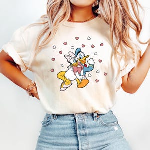 May include: A white t-shirt with a cartoon image of Donald Duck and Daisy Duck holding hands. The image is surrounded by pink and white hearts.
