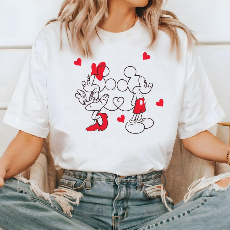 Op de afbeelding: Wit T-shirt met een afbeelding van Mickey Mouse en Minnie Mouse die hand in hand staan met rode hartjes eromheen.