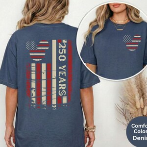 May include: A denim blue t-shirt with a distressed American flag design on the back, featuring a Mickey Mouse head shape and the text "250 YEARS". A smaller version of the flag design is on the front pocket.