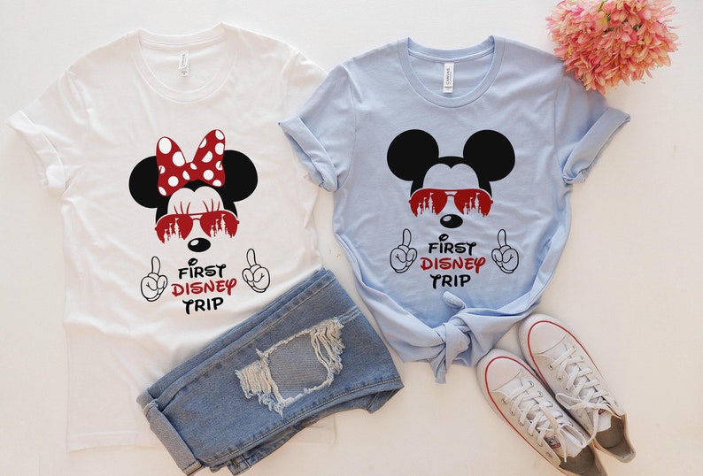 Op de afbeelding: Twee witte T-shirts met een Mickey Mouse en Minnie Mouse ontwerp. De shirts tonen de tekst "First Disney Trip" in rode letters. De shirts liggen op een wit oppervlak met een paar witte sneakers en een blauwe jeans.
