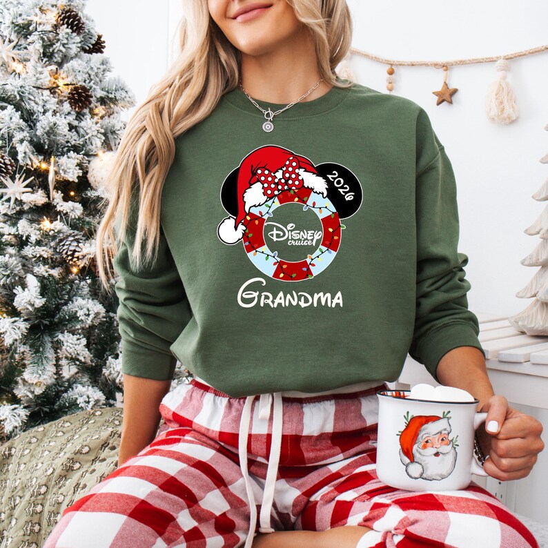 K&ouml;nnte beinhalten: Gr&uuml;nes Sweatshirt mit einem Disney Cruises-Motiv mit einem Mickey Mouse Weihnachtsmann-Hut, einer Rettungsboje und dem Wort "Oma". Das Jahr "2026" wird ebenfalls angezeigt. Eine Tasse mit einem Weihnachtsmann-Design wird gehalten.