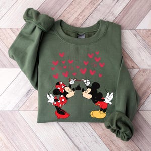 Puede incluir: Una sudadera verde oliva con Mickey y Minnie Mouse bes&aacute;ndose, con corazones rojos y la palabra "love" encima. Minnie lleva un lazo y un vestido de lunares rojos. Mickey lleva pantalones cortos rojos y zapatos amarillos.