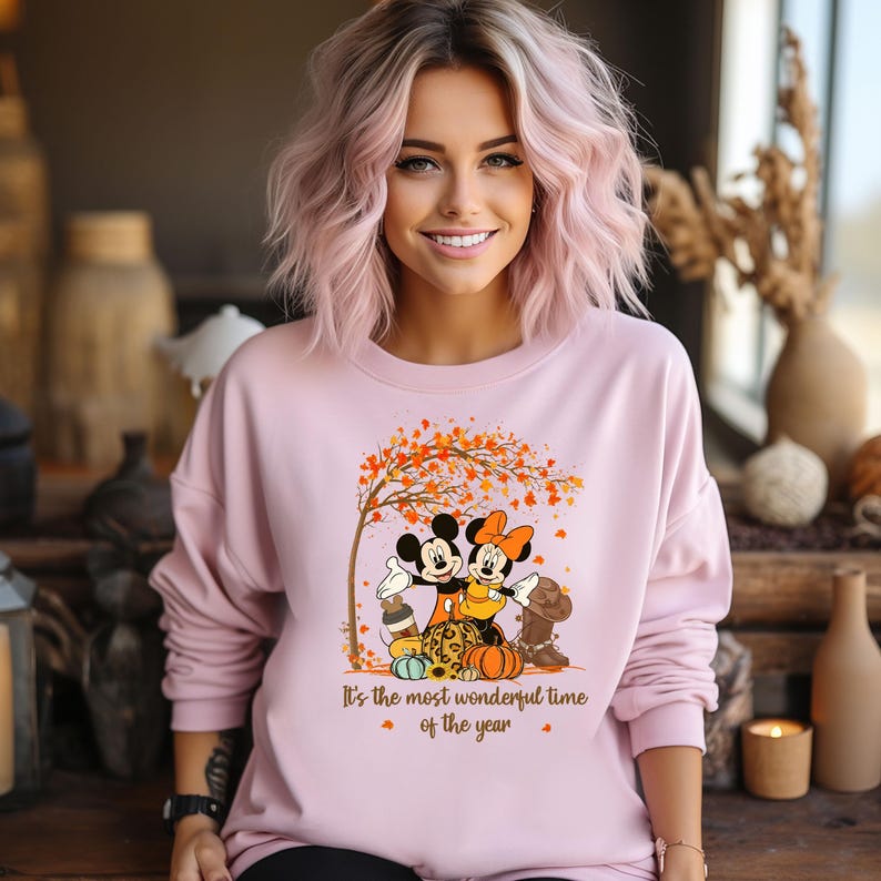 May include: A pale pink sweatshirt with a graphic of Mickey and Minnie Mouse beneath a tree with autumnal leaves. The text "It's the most wonderful time of the year" is below the graphic. The sweatshirt has long sleeves.