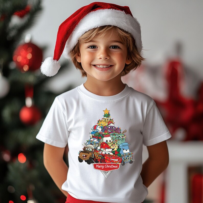 May include: A child wearing a white t-shirt featuring a Christmas tree design made of cartoon cars. The shirt has the text "Merry Christmas" on a red banner. The child is also wearing a red and white Santa hat.