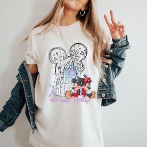 May include: Cream-coloured t-shirt with a Disney design. The graphic features a Mickey Mouse head outline, a castle, and Mickey and Minnie Mouse characters. The words "Disney Family" are written in a purple script.