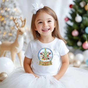 May include: A young girl wearing a white t-shirt with a snow globe design featuring Mickey and Minnie Mouse. The snow globe includes a Christmas tree and candy canes. The girl is smiling in front of a Christmas tree and decorations.