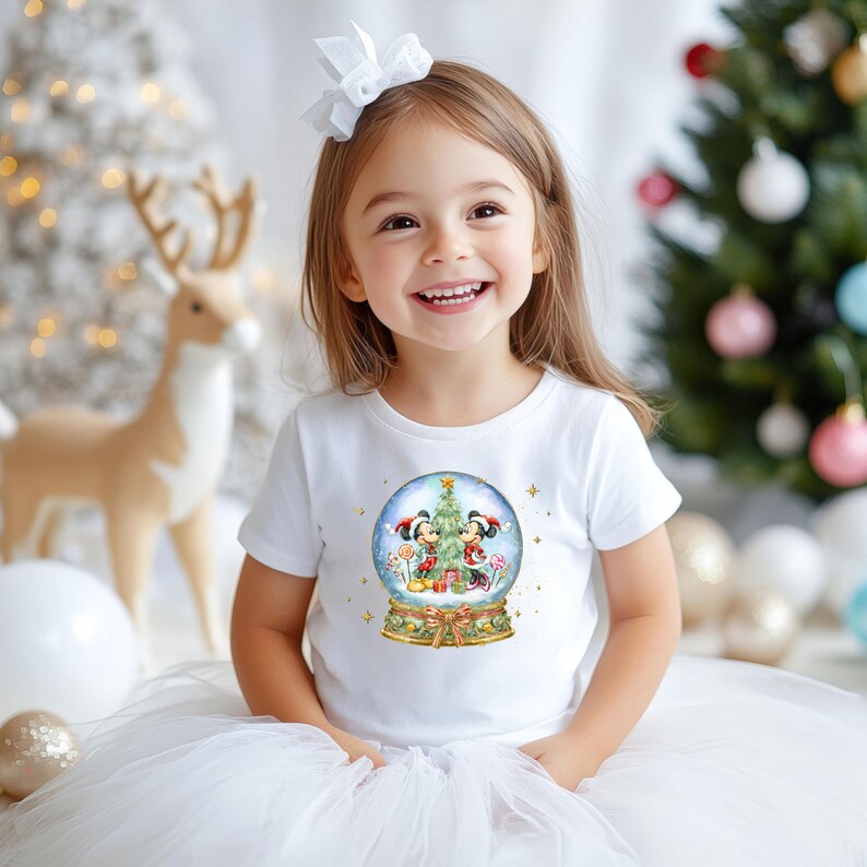 May include: A young girl wearing a white t-shirt with a snow globe design featuring Mickey and Minnie Mouse. The snow globe includes a Christmas tree and candy canes. The girl is smiling in front of a Christmas tree and decorations.