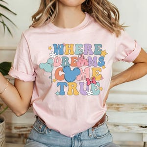 May include: Pink t-shirt with a graphic print that says "Where Dreams Come True" in colorful letters. The graphic also includes a Mickey Mouse head, a castle, stars, and balloons.