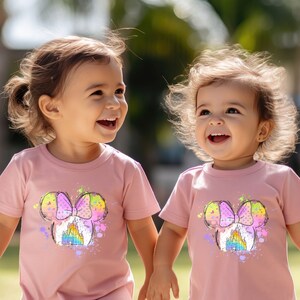Puede incluir: Dos ni&ntilde;as peque&ntilde;as con camisetas rosas con un gr&aacute;fico colorido de la cabeza de Mickey Mouse con un lazo y un castillo. Las ni&ntilde;as sonr&iacute;en y miran a la c&aacute;mara.