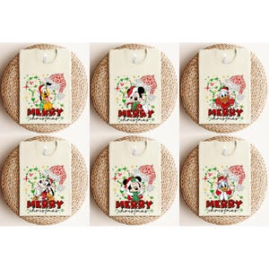 May include: Six cream-colored t-shirts with Christmas-themed designs featuring Disney characters. Each shirt displays a different character, including Mickey Mouse, Pluto, and Donald Duck, wearing Santa hats. The text "MERRY CHRISTMAS" is printed below each character.