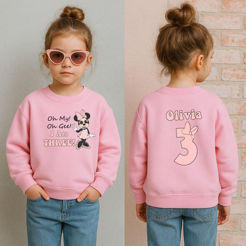 May include: Pink sweatshirt with a Minnie Mouse graphic and the text "Oh My! Oh Gee! I AM THREE!". The back of the sweatshirt has the name "Olivia" and the number "3". The sweatshirt is worn by a young child.