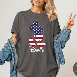 May include: Dark gray t-shirt featuring a Mickey Mouse silhouette filled with a stars and stripes design. The name "David" is written in white script below the character. The shirt is part of the Comfort Colors Pepper collection.