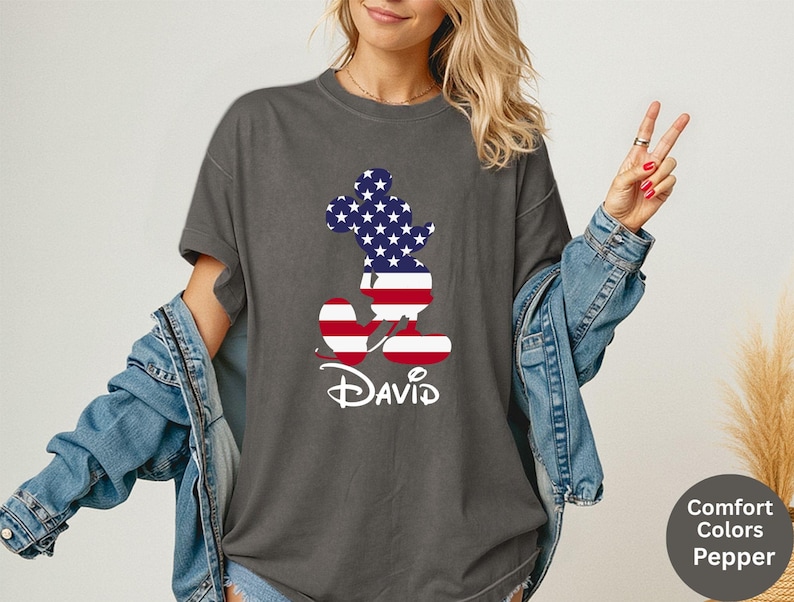 May include: Dark gray t-shirt featuring a Mickey Mouse silhouette filled with a stars and stripes design. The name "David" is written in white script below the character. The shirt is part of the Comfort Colors Pepper collection.