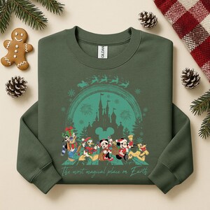 以下が含まれることがあります： ミッキーマウスとドナルドダックを含む、クリスマスの衣装を着たディズニーのキャラクターのターコイズ色のグラフィックが施された、濃い緑色のクルーネックのスウェットシャツ。お城の前。テキスト「The most magical place on Earth」が下にあります。