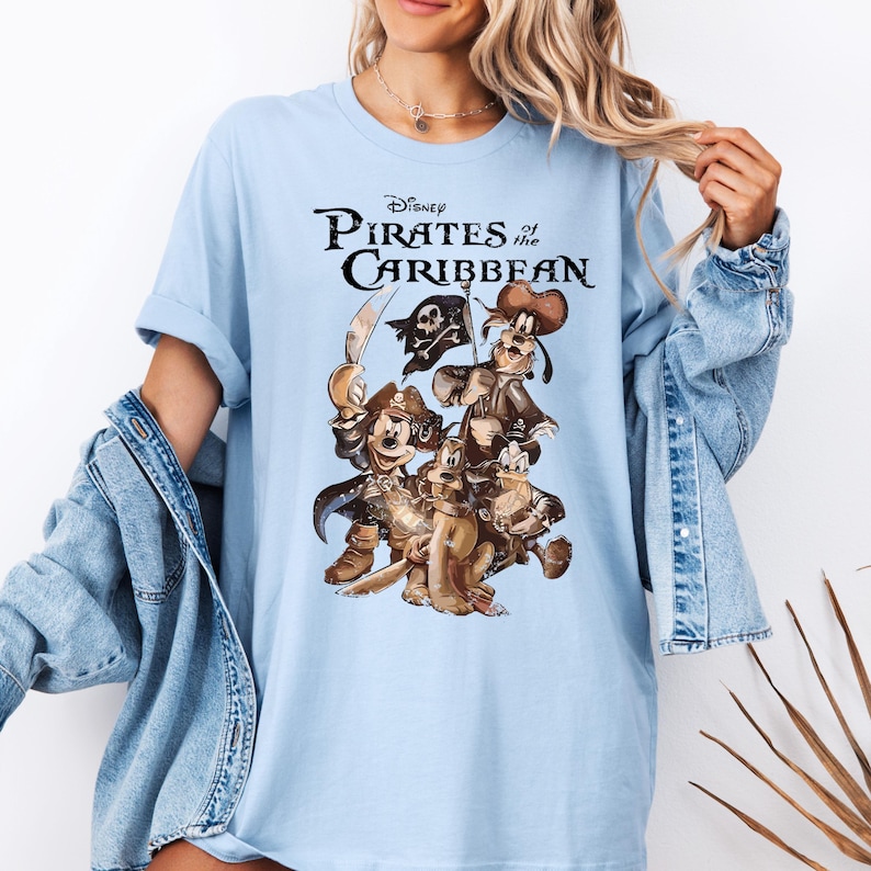 Pode incluir: Camiseta azul claro com uma estampa gr&aacute;fica de Mickey Mouse, Pateta e Donald Duck vestidos de piratas. O texto "Disney Pirates of the Caribbean" est&aacute; impresso acima dos personagens.