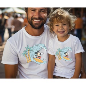 May include: White t-shirts featuring a graphic of Mickey Mouse surfing on a wave with palm trees. The shirts are for adults and children.