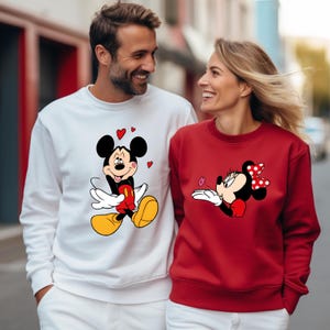May include: A couple wearing matching sweatshirts. One sweatshirt is white with a Mickey Mouse graphic and red hearts. The other is red with a Minnie Mouse graphic. Both characters are in a cartoon style.
